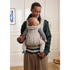 A man wearing a green and cream zip-up carrier jacket holds a baby in a beige quilted front baby carrier inside the jacket, standing in a brightly lit indoor room.
