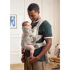 A man wearing a dark green t-shirt and khaki pants carries a baby in a light beige baby carrier vest. The baby is wearing a cream knit top and light pink pants.