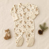 A baby footed sleeper laid flat on a light fabric background, featuring an allover deer print in tan and brown on an off-white base, with textured cream footies and a snap closure at the neck. A wooden fox toy, pine branch, and pinecone are styled around it.