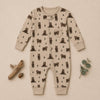 A baby long-sleeve footless romper in beige with a repeating print of dark brown bears and small pine trees, styled flat-lay on a beige background with decorative wood and eucalyptus.
