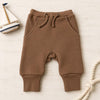 A pair of baby jogger pants in a warm brown color, styled flat-lay on a light wood background with a small toy sailboat and a piece of rope.