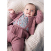 A baby lying on a textured cream quilted blanket, wearing a dusty rose knit cardigan and matching corduroy-style overalls over a floral print bodysuit.
